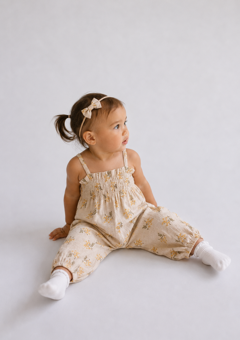 A toddler girl sitting on a light gray studio floor wearing a cream-colored floral romper with thin shoulder straps, white socks, and a matching bow headband.
