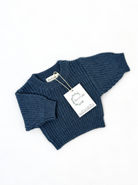 A dusty sapphire ribbed knit sweater laid flat on a white background with a white paper brand tag attached.